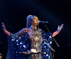 A singer wearing a shiny, reflective outfit performs on stage with arms outstretched at one of the vibrant LATAM music festivals, standing behind a keyboard and singing into a microphone.